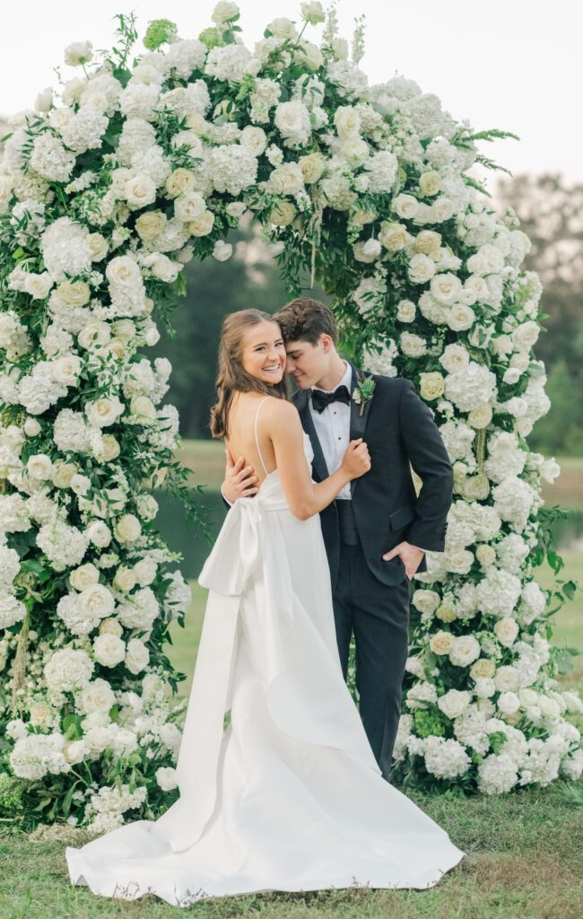 bride and groom in nuzzle pose for a portrait with bride turned looking over her shoulder to show off the bow on the back of her dress. their floral arch of the ceremony is the backdrop