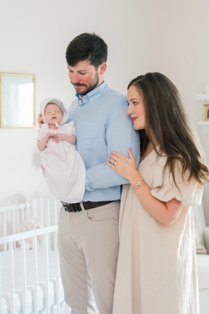 mom and dad standing in nursery dad is holding baby and mom is linked to dads side both looking and smiling and baby