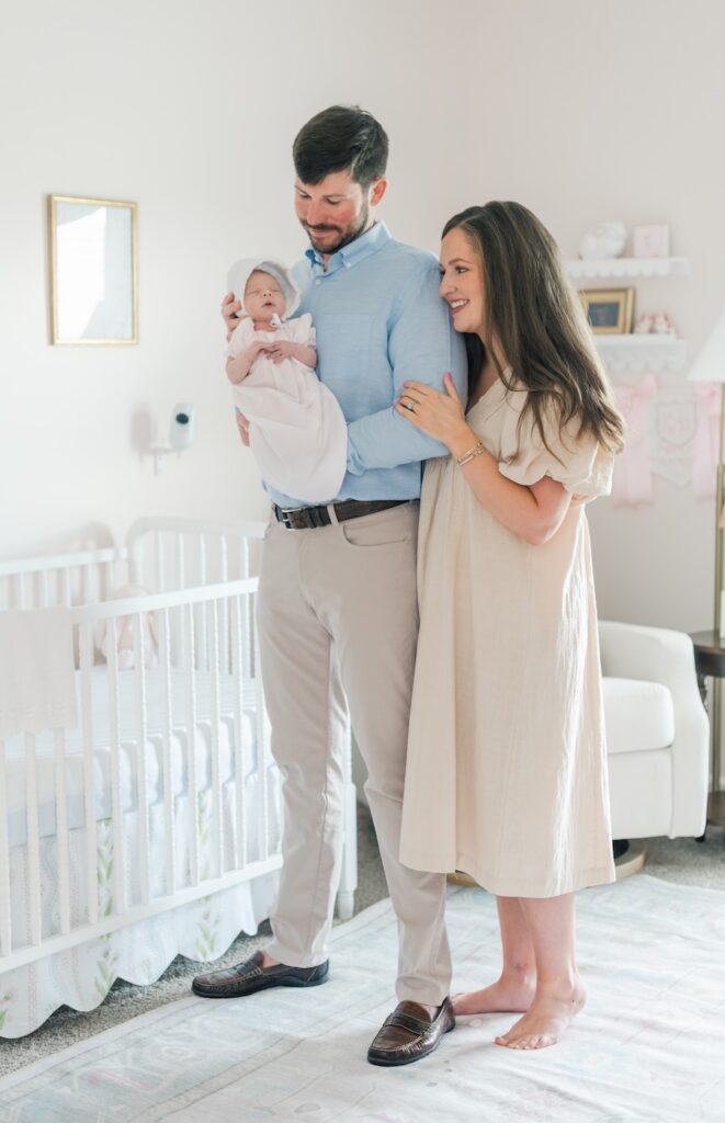 mom and dad standing in nursery dad is holding baby and mom is linked to dads side both looking and smiling and baby