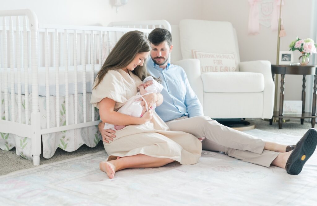 a new mom and dad sit on the floor in nursery looking down at baby as they are propped against crib