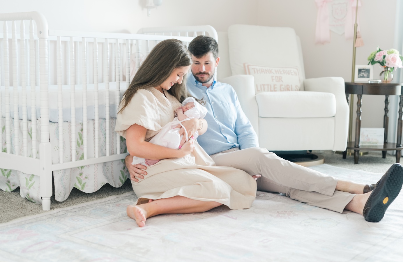 a new mom and dad sit on the floor in nursery looking down at baby as they are propped against crib