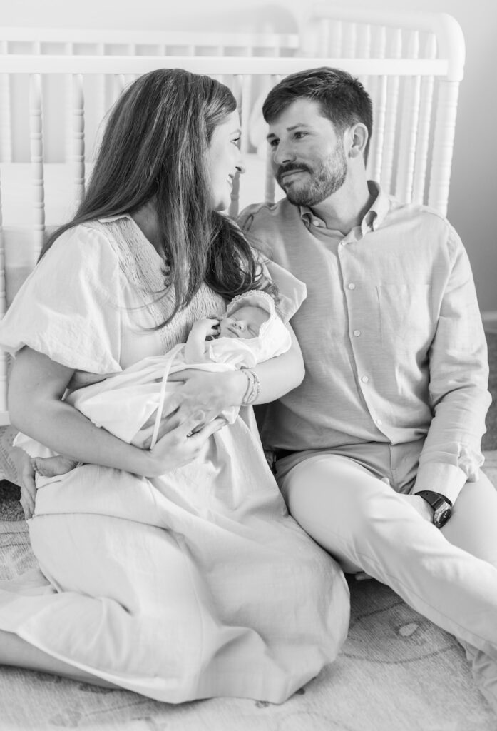 black and white photo mom and dad sitting on the floor in front of the crib holding baby 